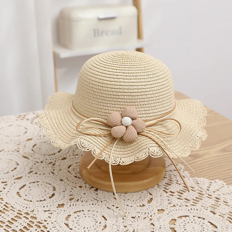 A charming beige straw hat featuring a scalloped brim and a delicate flower decoration made of natural-toned fabric and wooden beads, tied with a simple bow. Perfect for a stylish and elegant summer look for children.