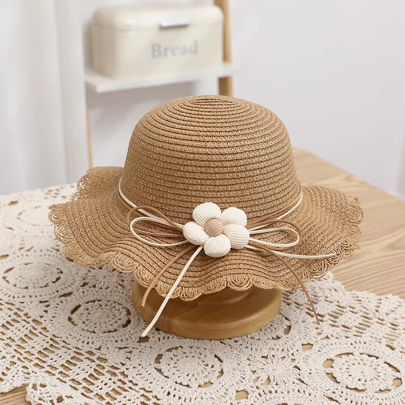 A classic brown straw hat featuring a scalloped brim and a cream-colored fabric flower with wooden bead details, tied with a natural-toned bow. Perfect for a timeless and elegant summer look for children.