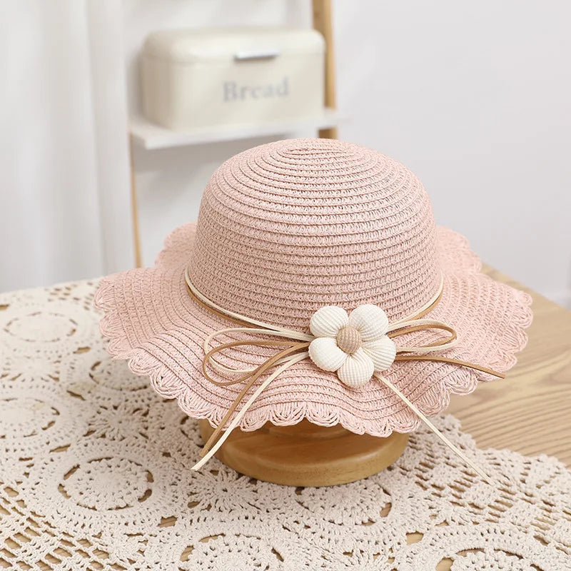 A lovely pink straw hat featuring a scalloped brim and a delicate cream-colored flower decoration made of fabric and wooden beads, tied with a matching bow. Ideal for a charming and elegant summer look for children.