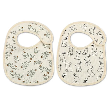 A set of two baby bibs made of soft fabric. One bib features a botanical pattern with green leaves and small brown berries, while the other has a playful design of outlined bunny illustrations. Both bibs have a neutral beige background and a rounded shape with a side closure for comfort.