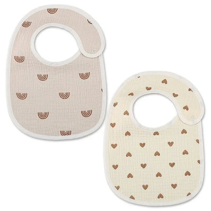 A set of two baby bibs made of soft fabric. One bib features a pattern of small brown rainbows on a beige background, while the other displays a design of tiny brown hearts on a light cream background. Both bibs have a rounded shape and a side closure for easy wear.