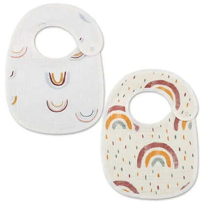 A set of two baby bibs made of soft fabric. One bib features small minimalistic rainbows in warm tones on a beige background, while the other displays larger, vibrant rainbows with scattered colorful dots. Both bibs have a rounded shape and a side closure for easy use.