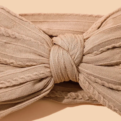 Extreme close-up of beige cotton bow headband focusing on the intricate center knot construction, showcasing the textured ribbed fabric pattern and symmetrical pleating detail against a warm beige background