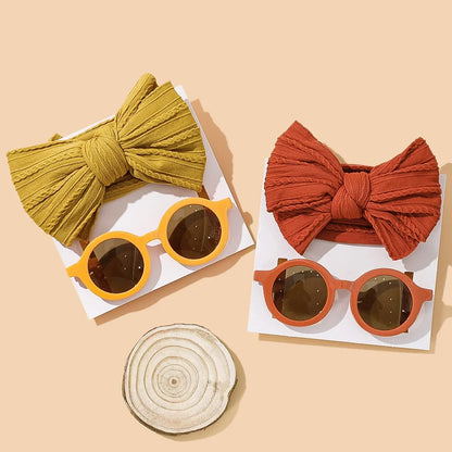 Stylish autumn baby accessories collection featuring two coordinating sets: a mustard yellow bow with matching round sunglasses and a terracotta bow with matching round sunglasses, artfully arranged with a natural wood coaster on warm beige background