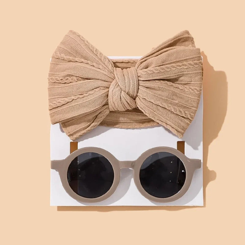 Stylish baby accessory set featuring a textured beige cotton bow headband paired with round gray sunglasses with dark lenses, arranged on white card against warm beige background for a modern minimalist aesthetic