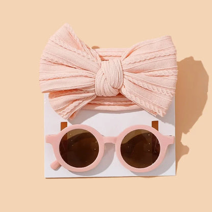 Delicate baby accessory set featuring a textured blush pink cotton bow headband paired with matching round blush sunglasses with brown lenses, elegantly presented on white card against warm beige background for a soft, feminine aesthetic