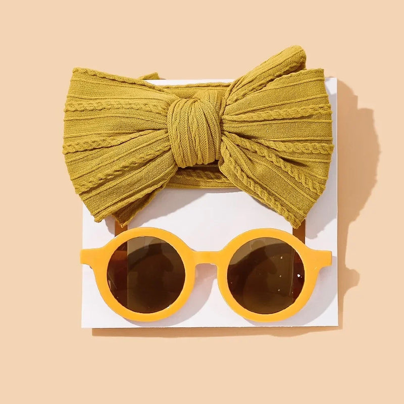 Trendy baby accessory set featuring a textured mustard yellow cotton bow headband paired with matching round mustard sunglasses with brown lenses, artfully displayed on white card against warm beige background for a modern autumn-inspired look