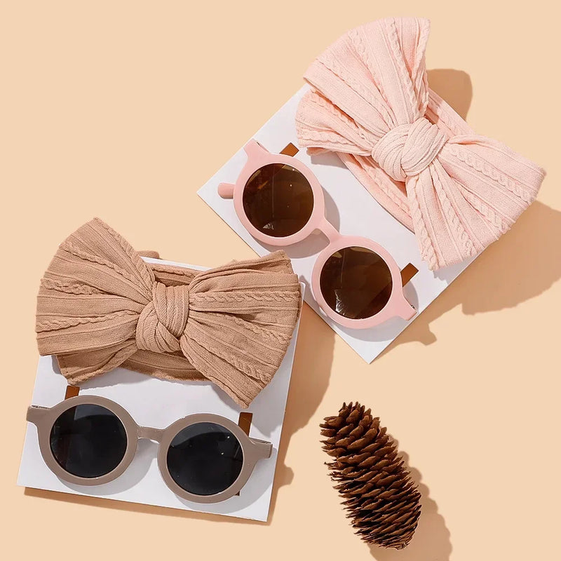 Elegant neutral baby accessories collection featuring two coordinating sets: a beige cotton bow with matching round sunglasses and a blush pink bow with matching round sunglasses, artfully styled with a decorative pinecone on warm beige background