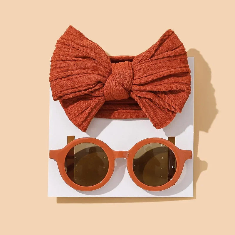 Coordinated baby accessory set featuring a textured terracotta cotton bow headband paired with matching round terracotta sunglasses with brown lenses, displayed on white card against warm beige background for a modern earth-toned look