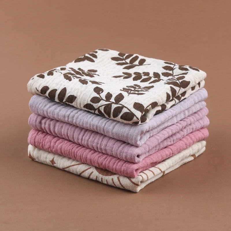 Elegant stack of folded baby towels featuring botanical print in cream and brown on top, followed by soft lavender, dusty rose, and natural cream colors, showcasing the complete collection's coordinated palette on warm brown background