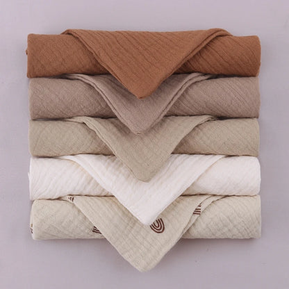 Neatly folded stack of baby towels in a gradient of earth tones, from rich brown at top through taupe, beige, pure white, to patterned cream at bottom, showcasing the complete neutral collection's sophisticated color palette on soft lilac background