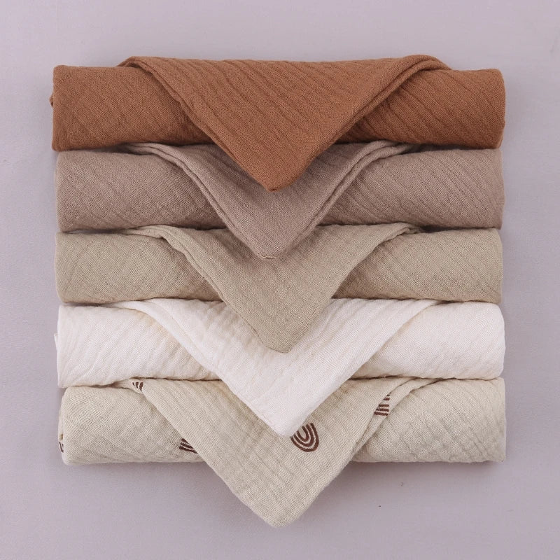 Neatly folded stack of baby towels in a gradient of earth tones, from rich brown at top through taupe, beige, pure white, to patterned cream at bottom, showcasing the complete neutral collection's sophisticated color palette on soft lilac background