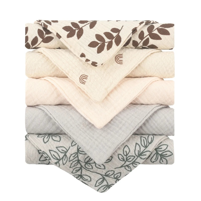 Collection of quilted baby towels featuring cream towels with brown leaf patterns and rainbow motifs, solid cream towels, and grey towels with delicate botanical line drawings, all with diamond-textured cotton for gentle baby care