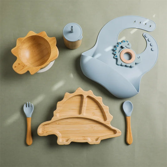 Blue dinosaur-themed bamboo toddler dining set featuring a silicone bib, dinosaur-shaped divided plate, bamboo bowl with suction base, sippy cup, spoon, fork, and a matching dinosaur-shaped teether, designed for safe and enjoyable mealtime experiences.