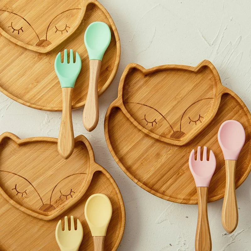 Set of bamboo fox-shaped baby plates with divided sections, each paired with wooden spoons and forks featuring colorful silicone tips in green, yellow, and pink, designed for safe and enjoyable toddler meals.