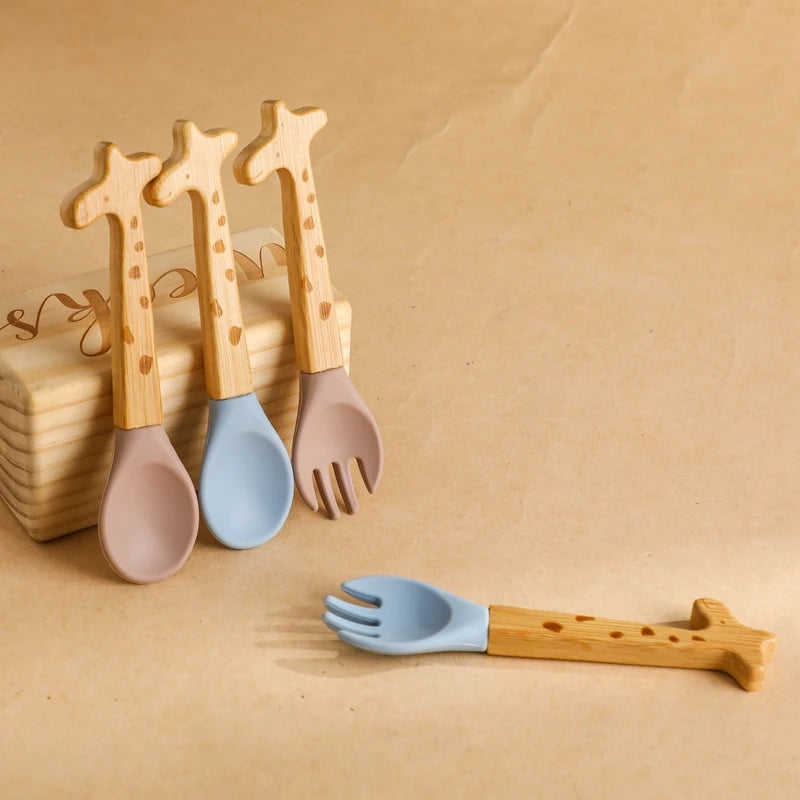 A set of bamboo cutlery with giraffe-shaped handles, including spoons and forks with silicone tips in soft pastel colors such as blue and taupe, designed for children.