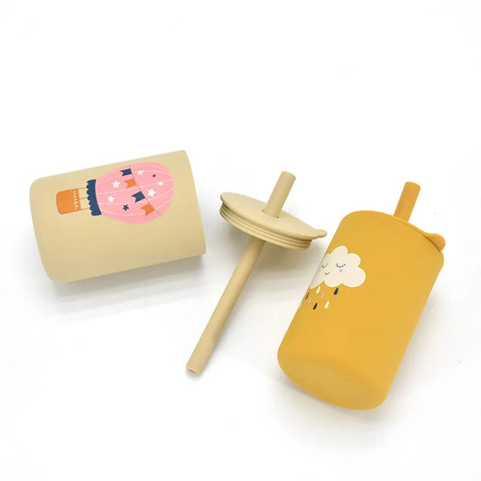 Collection of silicone baby straw cups in beige and yellow tones, featuring whimsical designs including a hot air balloon and sleeping cloud, with matching wooden-look straw lids for safe and stylish toddler drinking