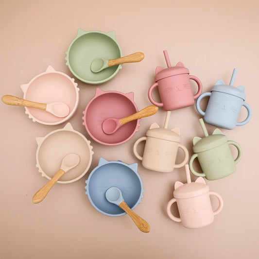 Artistic flatlay of complete silicone baby feeding collection featuring scalloped bowls and matching sippy cups in soft pastel colors - blush pink, sage green, powder blue, cream, and dusty rose - each paired with wooden spoons, arranged in a circular pattern on neutral background