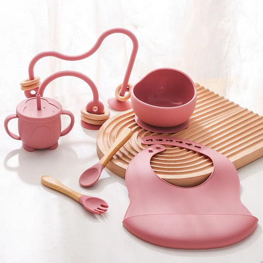 Pink silicone baby feeding set featuring a bib, suction bowl, sippy cup with handles, spoon, fork, and a bead maze toy, arranged on a wooden textured mat, designed for mealtime convenience and sensory play.