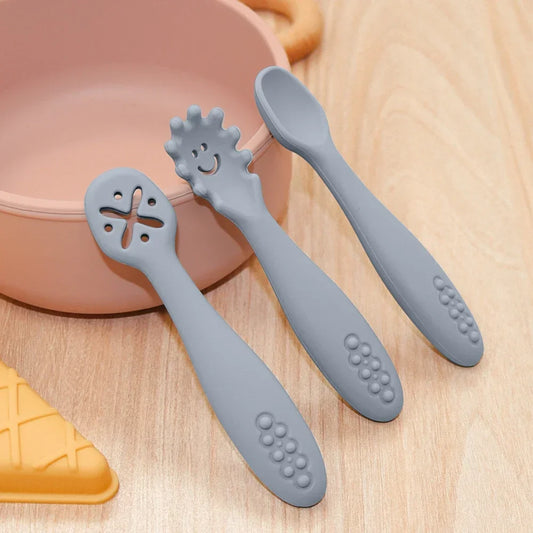 Lifestyle image of grey silicone baby spoons displayed on wooden surface next to pink and yellow baby dishes, featuring three spoon designs with smooth, cross-pattern, and smiling sun textures, showcasing their practical use in a mealtime setting