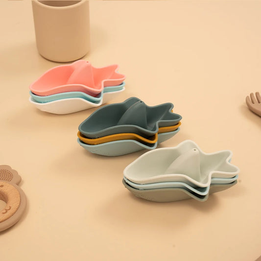 Three stacks of silicone bath boats in coordinating colors - coral pink with mint and cream, forest green with sage and mustard, and neutral cream with soft sage tones, featuring a modern leaf-shaped design for engaging bathtime play