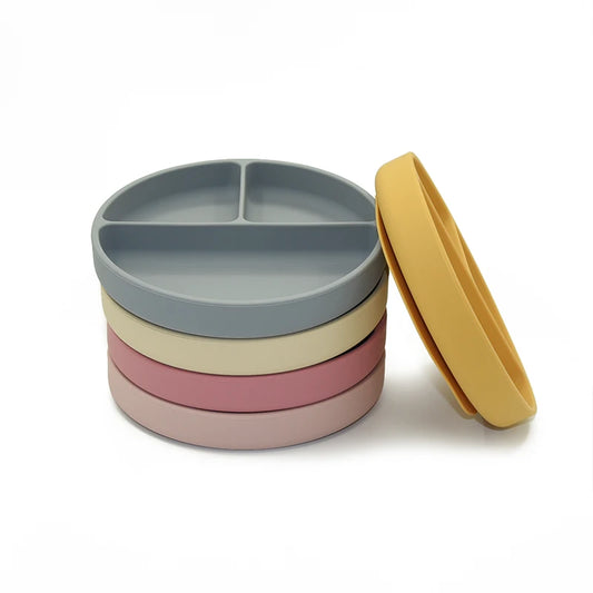 A stack of multicolored silicone divided plates in shades of gray, beige, pink, and mustard yellow. Each plate features three compartments, designed for portion control and easy toddler feeding, made from food-grade silicone for durability and easy cleaning.