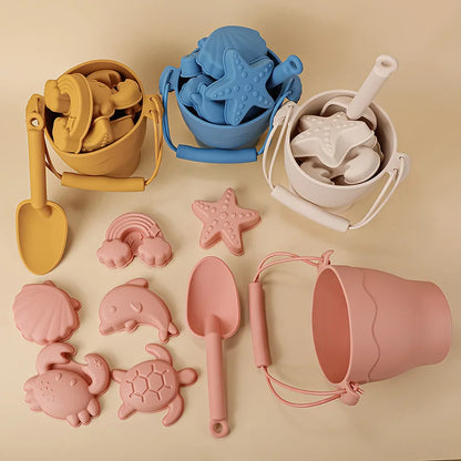 Artistic flat lay of silicone beach toy collection featuring buckets with handles in mustard, blue, and cream, alongside coral pink set with matching scoops and marine-themed molds including starfish, dolphins, turtles, and crabs, arranged on warm beige background