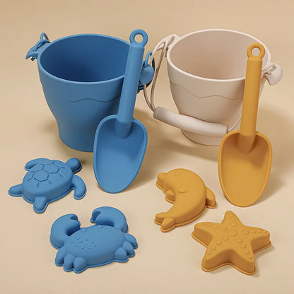 Side-by-side comparison of silicone beach toy sets in dusty blue and mustard yellow, each featuring a bucket with handle, scoop, and sea creature molds including turtle, crab, and starfish, showcasing the modern color options on a neutral beige background