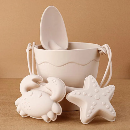 Close-up of cream silicone beach toys featuring a textured bucket with handle and scoop, alongside detailed crab and starfish molds with raised patterns, showcasing the high-quality finish and modern neutral aesthetic on a warm beige surface