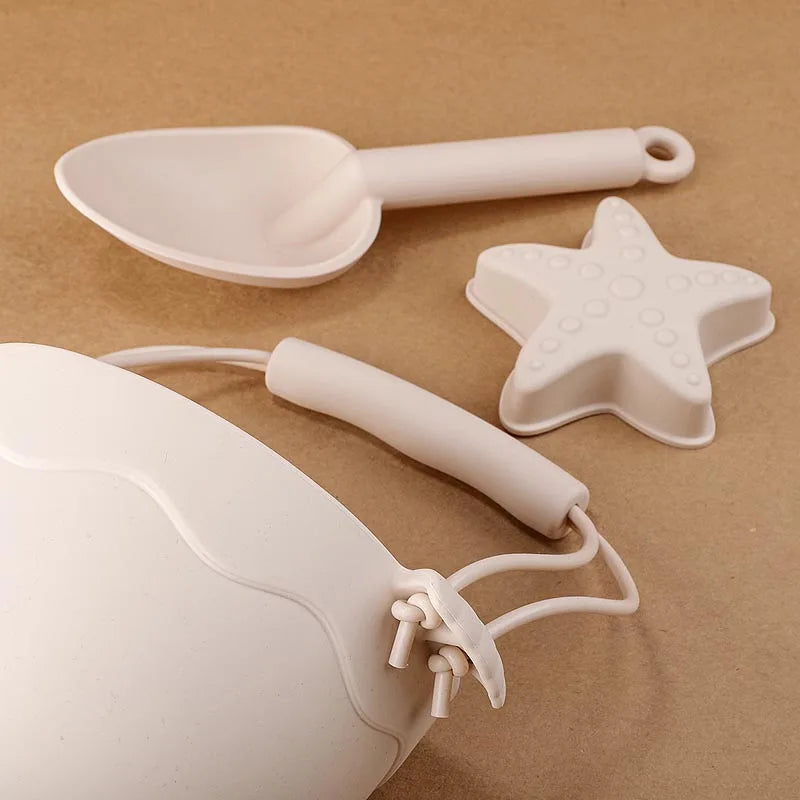 Artistic composition of cream silicone beach toys highlighting key features: ergonomic scoop handle, starfish mold texture, and durable bucket strap design, arranged on warm beige background to showcase the modern, minimalist aesthetic