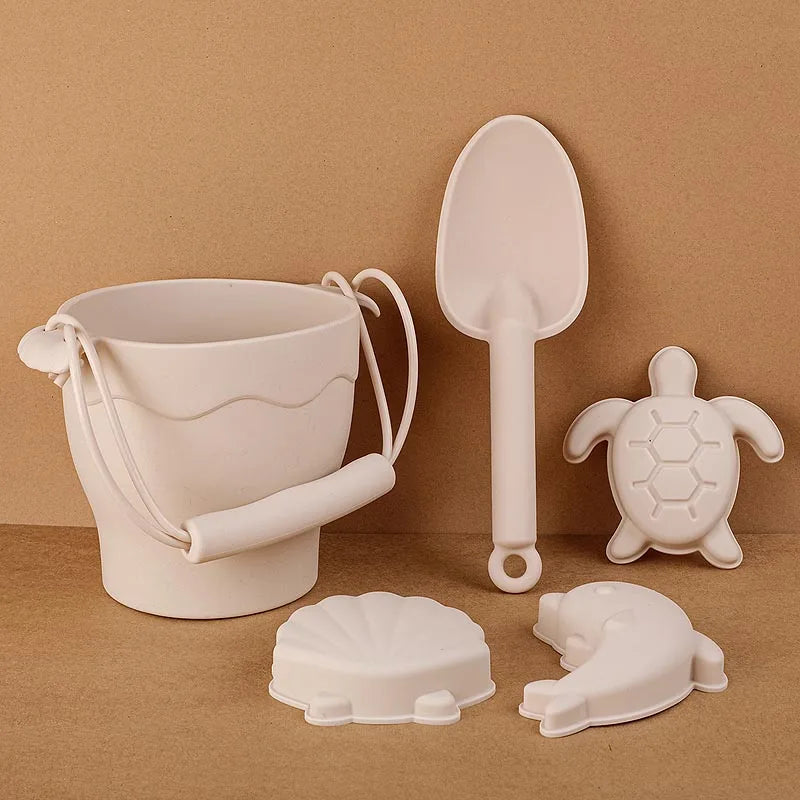 Minimalist cream silicone beach toy set styled on warm beige background, featuring bucket with handle, large scoop, and sea creature molds including turtle and dolphin shapes, showcasing the modern aesthetic of eco-friendly beach play