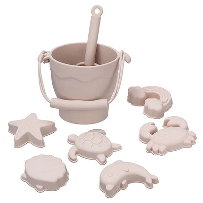 Complete cream-colored silicone beach toy set featuring a bucket with handle and scoop, plus six marine-themed molds including turtle, starfish, shell, rainbow, crab, and dolphin shapes, designed in a soothing neutral tone for modern sensory play