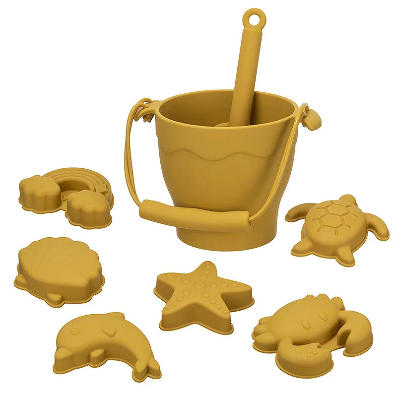 Complete mustard yellow silicone beach toy set featuring a bucket with handle and scoop, plus six ocean-themed molds including turtle, starfish, shell, rainbow, crab, and dolphin shapes, perfect for modern sensory play in a trendy warm tone