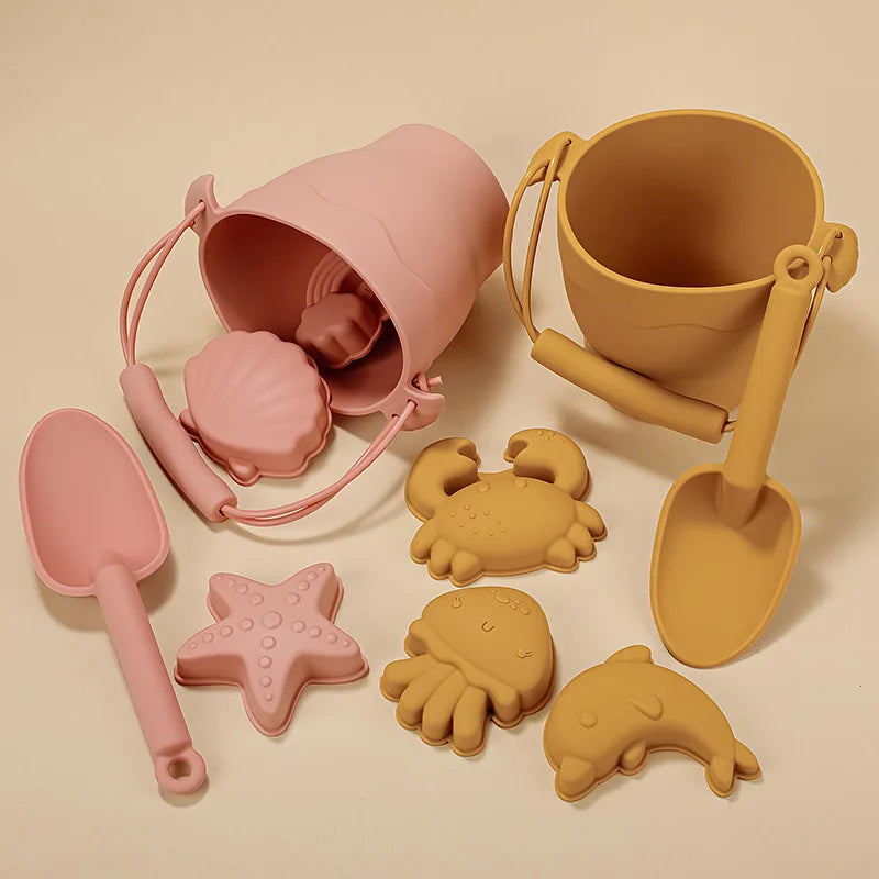 Coordinating silicone beach toy sets in coral pink and warm mustard, each featuring a bucket with handle, scoop, and sea creature molds including starfish, crab, and dolphin shapes, artfully arranged on a neutral beige surface