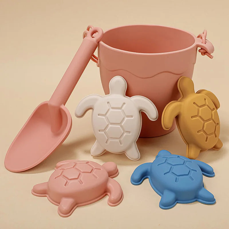 Silicone beach toy set featuring coral pink bucket and scoop with matching turtle mold, accompanied by coordinating turtle molds in cream, mustard, and dusty blue, displaying the full color range on a warm beige background