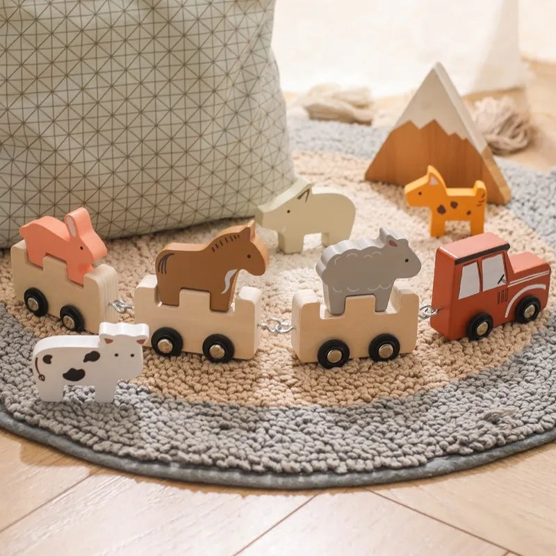 Wooden animal train set arranged on grey circular rug with geometric cushion backdrop and wooden mountain peak, featuring a red locomotive leading natural wood carriages with safari animals including coral fox, brown horse, grey elephant, and spotted dalmatian figure, creating a cozy Nordic-inspired play setting