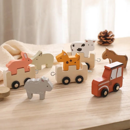 Wooden animal train set photographed on light wooden surface with soft fabric backdrop and natural pinecone accent, featuring a red locomotive leading interconnected carriages with safari animals including coral fox, beige polar bear, orange giraffe, spotted cow, grey elephant, and brown horse figures in a minimalist Montessori-style arrangement