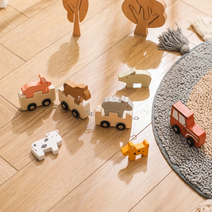 Wooden animal train set arranged on light wooden flooring with grey round rug and decorative wooden trees, featuring interconnected cars with safari animals including fox, bear, elephant, and giraffe figures, accompanied by a red locomotive engine creating a charming play environment