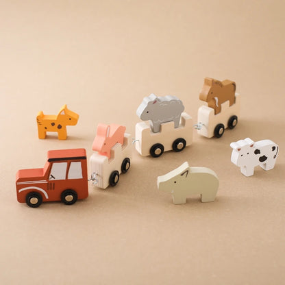 Montessori-style wooden safari animal train set featuring a red locomotive leading interconnected natural wood carriages, each carrying unique animal figures including orange giraffe, coral fox, grey elephant, and brown horse, with additional spotted cow and polar bear figures, arranged against a warm beige background for a clean product display