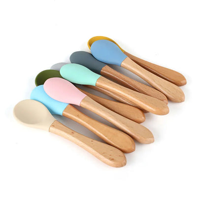 Collection of wooden baby spoons with pastel-colored silicone tips including mint, pink, blue, beige, olive green, and grey, each featuring natural wooden handles and soft silicone bowls for gentle infant feeding