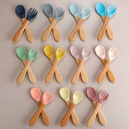 Complete collection of wooden baby utensils featuring spoon and fork sets in eleven colors ranging from navy blue, sky blue, turquoise, mint green, yellow, to various pastel shades, each combining natural wooden handles with soft silicone tips for safe infant feeding