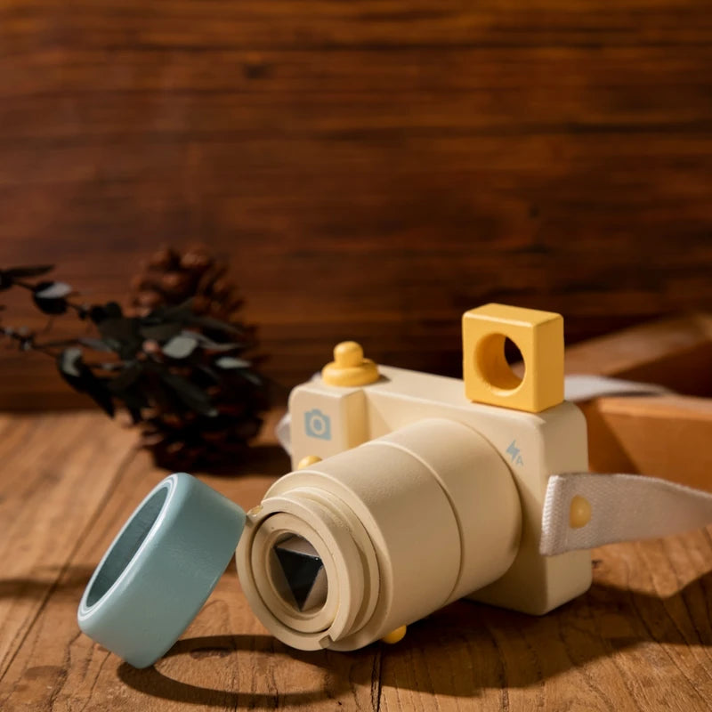 Handcrafted wooden toy camera in cream and mint colors displayed on rustic wooden table with dried eucalyptus accent, featuring interchangeable lens blocks and geometric design elements for imaginative play and fine motor development