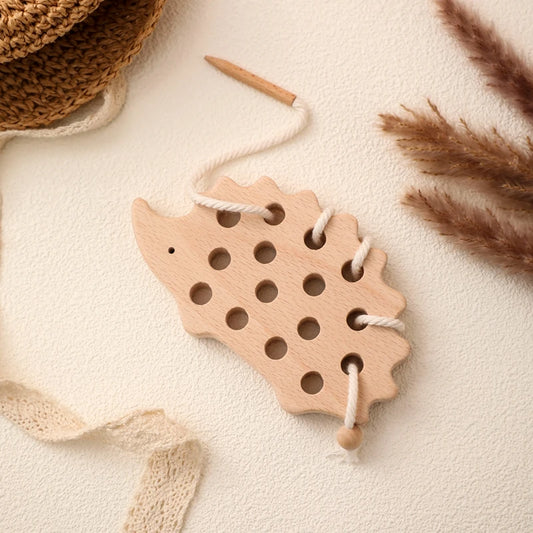 Terracotta wooden hedgehog threading toy with natural cotton lacing cord, styled with dried pampas grass and woven basket elements on cream textured surface, showcasing the eco-friendly Montessori learning toy in a modern minimalist setting