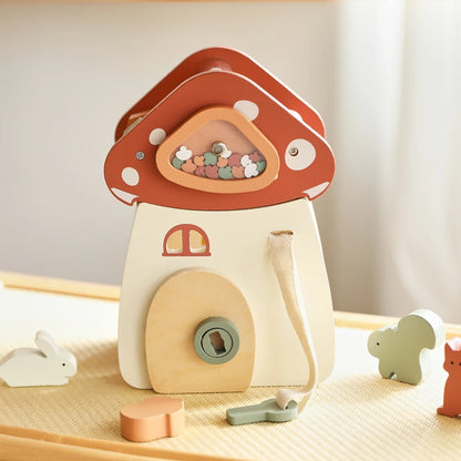 Front-facing view of wooden mushroom dollhouse with terracotta red cap adorned with white polka dots and sparkling gemstone window, featuring a cream-colored base with rounded doorway and sage green keyhole, surrounded by miniature wooden woodland creatures on a textured cream surface