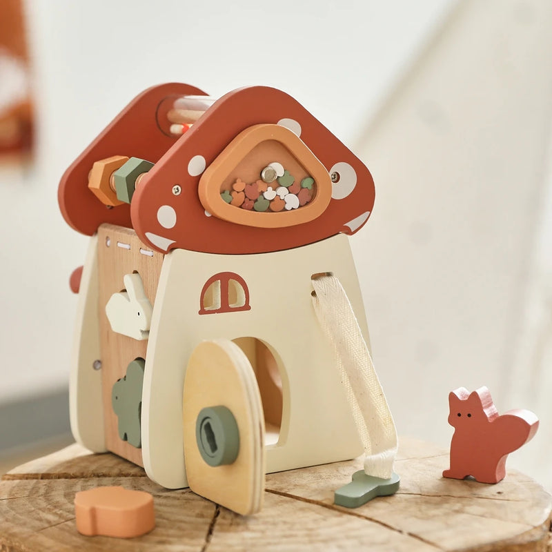 Whimsical wooden mushroom house playset featuring a cream-colored base with terracotta red caps, decorated with small round windows and doors, accompanied by miniature wooden accessories including a red fox figure, displayed on rustic wooden surface creating a magical woodland scene