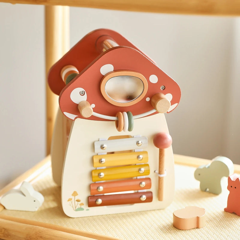 Enchanting wooden mushroom house with built-in rainbow xylophone bars in warm yellow to terracotta tones, photographed in golden sunlight that highlights the cream-colored base, red polka-dotted cap, and delicate details, with woodland animal figures creating a magical play scene