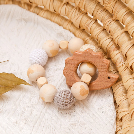 Natural wooden teething bracelet with terracotta-colored hedgehog pendant displayed on white muslin fabric against woven basket backdrop, featuring alternating smooth beech wood beads and grey crochet cotton spheres, styled with dried autumn leaves for an organic nursery aesthetic