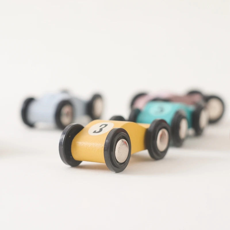 Close-up of a mustard yellow wooden toy car with number 3, featuring crisp focus on its streamlined design and black and white wheels, while additional pastel-colored cars create a soft, artistic blur in the background